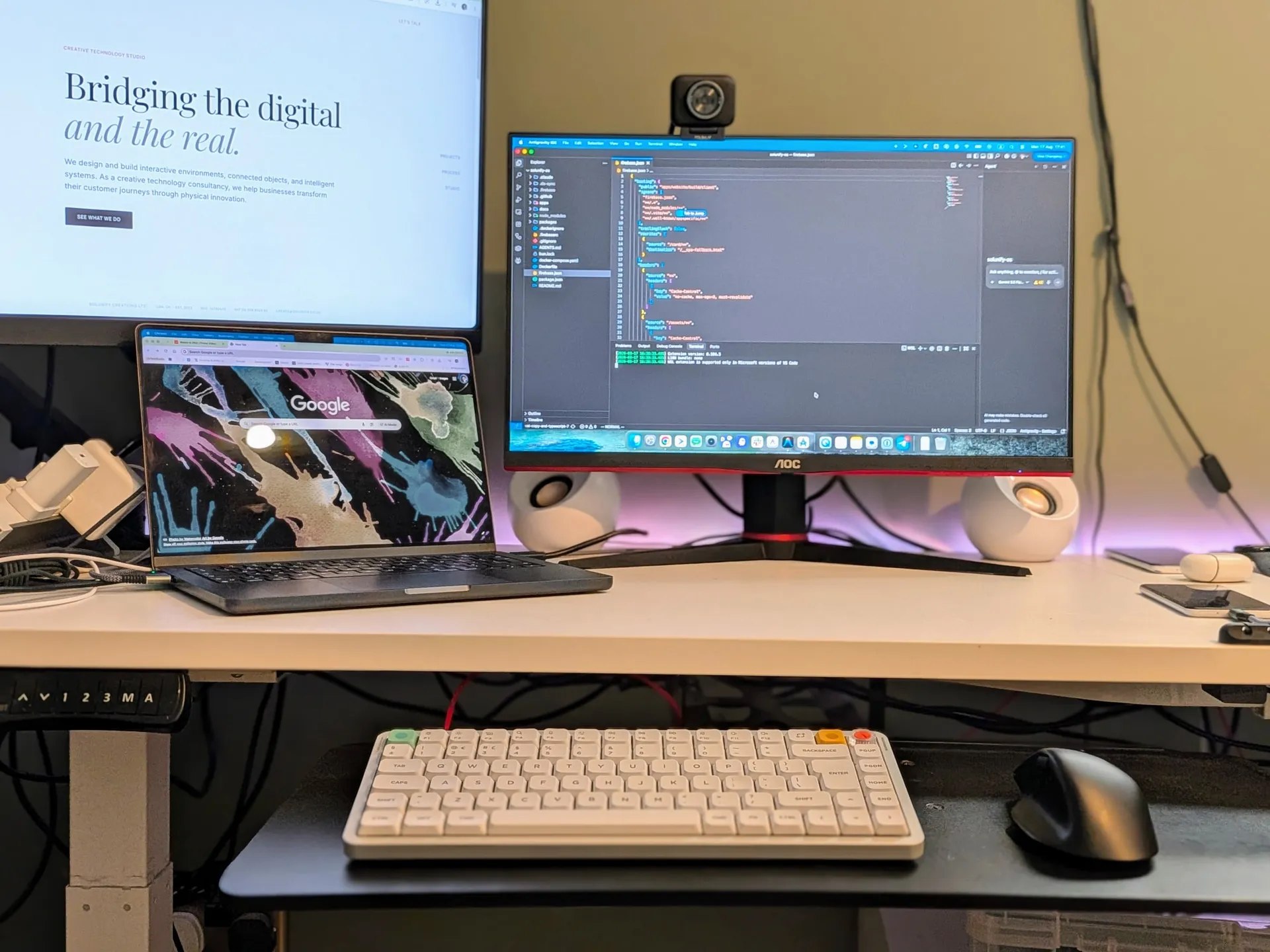 The software desk: a standing desk with a monitor showing the studio's own codebase in an editor, alongside a laptop and a mechanical keyboard
